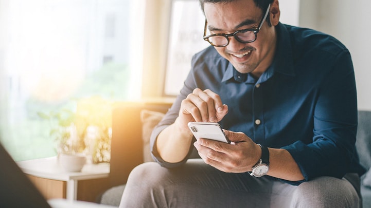A man using a smartphone while sitting on a couch and smiling.